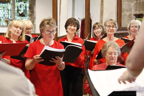 The image shows several singers in the choir performing at a concert. They are wearing choir uniform of red tops and black trousers and singing from black music folders. They are looking towards their Musical Director who is just out of shot.