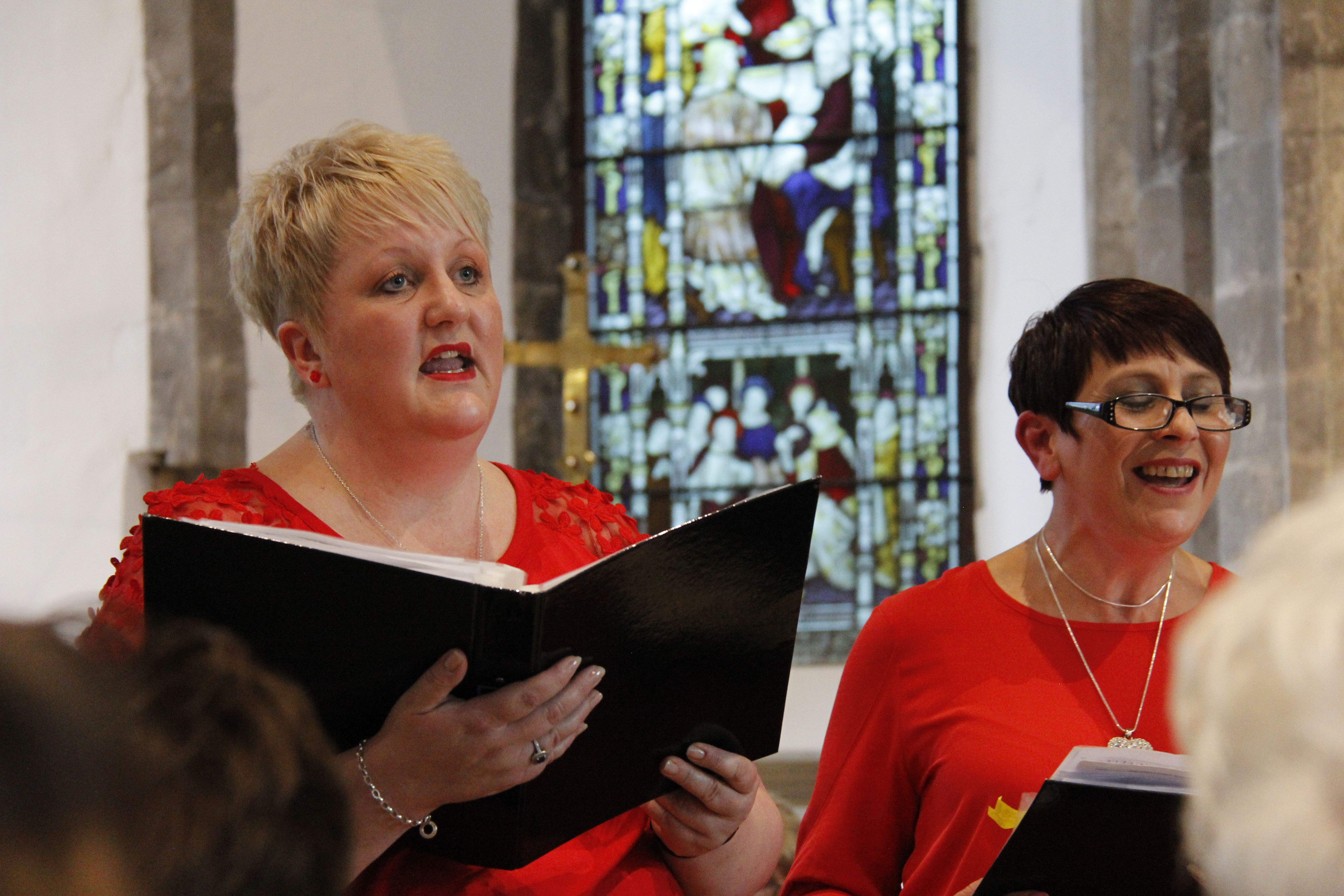 Two singers stand side by side singing from their music folders. The singer on the left is taller with short, blond hair and the singer on the right is shorter, with short brown hair and glasses. They're both wearing red tops and their expressions are joyful.
